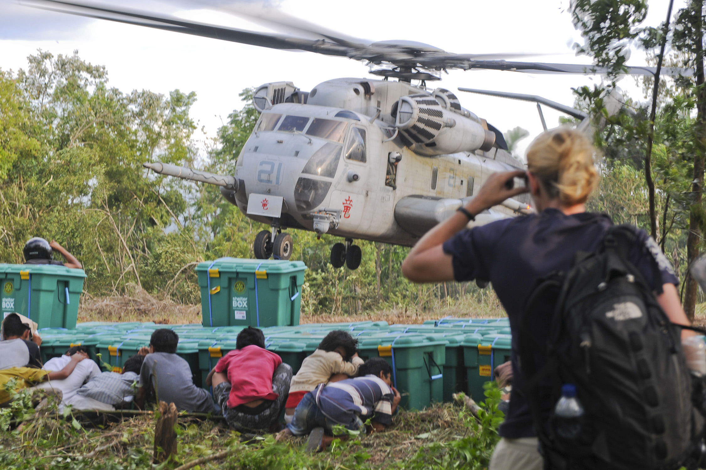 Children taking shelter behind a stack of Shelter boxes as a helicopter lands